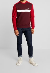 Uomo che indossa un felpa bordeaux e rossa con una striscia bianca, jeans scuri arrotolati e sneakers bianche alte, in piedi su uno sfondo semplice.