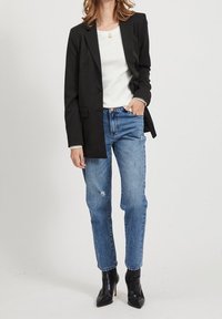 Blazer noir ajusté porté sur un pull blanc, associé à un jean bleu clair déchiré et des bottines à talons noires. Textures de tissu lisses.