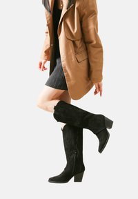 Botas negras hasta la rodilla de ante con detalles texturizados, tacones de bloque y cremalleras en los laterales; combinadas con un abrigo marrón y un vestido negro.