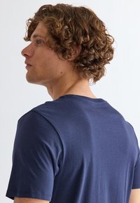 Mand med krøllet brunt hår iført en mørkeblå kortærmet t-shirt, vendt mod venstre mod en ensfarvet lys baggrund.