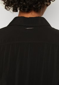 Čierna, ľahká košeľa s špicatým golierom a jemným leskom. Má malú kovovú značku na zadnej strane a voľný strih.