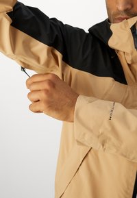 Veste légère beige et noire avec manches longues, capuche, poignets ajustables et détails de logo sur la manche.