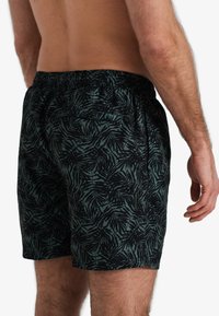 Zwarte en groene gestreepte zwemshorts met een elastische tailleband en een achterzak, voorzien van een palmbladontwerp op een lichte stof.