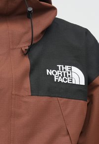Καφέ και μαύρο μπουφάν με κουκούλα και φερμουάρ σε τσέπη, με υφή υφάσματος και το λογότυπο The North Face κεντημένο σε λευκό χρώμα.