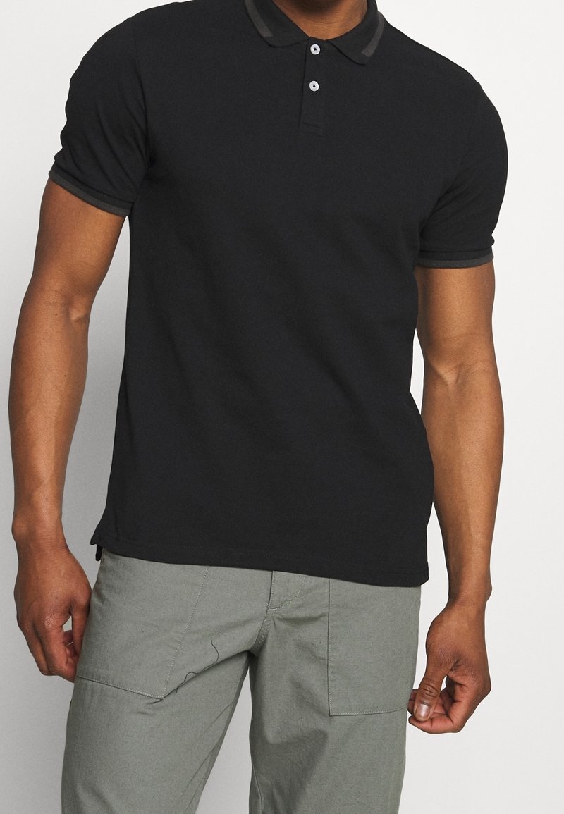 Homme portant un polo noir à manches courtes avec deux boutons et un pantalon gris clair, debout devant un fond uni.