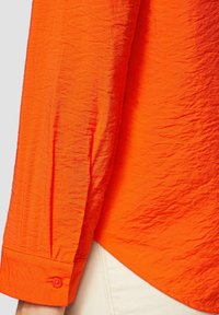 Chemise orange vif avec une texture froissée, des poignets à boutons et un ourlet arrondi. Le tissu semble léger et décontracté.