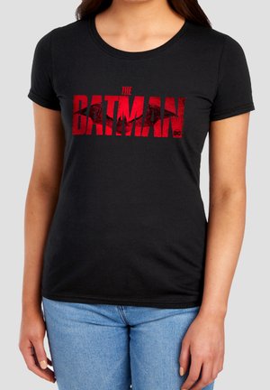 Frau trägt ein schwarzes T-Shirt mit rotem "The Batman"-Text und kleinem DC-Logo, kombiniert mit hellblauen Jeans, vor grauem Hintergrund stehend.
