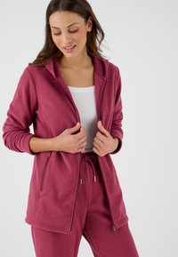 Giacca in pile rosa con zip e cappuccio, tasche frontali e polsini a coste, abbinata a pantaloni coordinati. Texture morbida e design casual.
