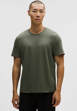 Mand med kort hår iført en olivengrøn T-shirt med korte ærmer og sorte bukser, stående foran en ensfarvet lysegrå baggrund.
