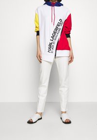 Bunter Hoodie mit blauen, gelben, roten und weißen Abschnitten, mit einem diagonalen Reißverschluss und schwarzem Text. Kombiniert mit weißen Hosen und schwarzen Sandalen.