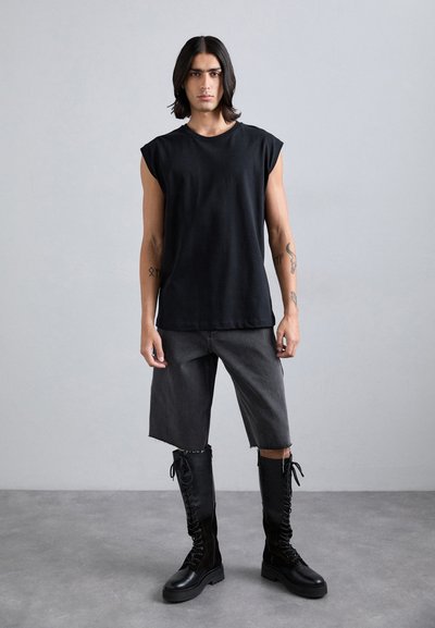 Zwarte mouwloze t-shirt, grijze denim shorts, zwarte kniehoge veterschoenen. Model met lang haar staat tegen een neutrale achtergrond.