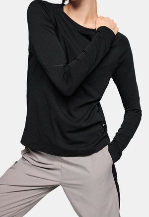 Personne portant un haut à manches longues noir et un pantalon gris clair, posant avec une main sur l'épaule et l'autre dans la poche.