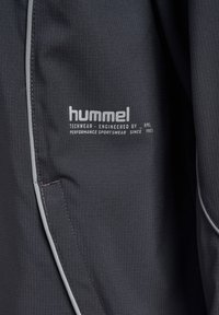 Donkergrijze stof met wit "hummel"-logo en tekst "Techwear engineered by HML, performance sportswear since 1923" nabij een verticale naad.