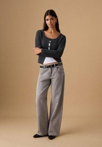 Grau gerippter Cardigan mit drei Knöpfen, kombiniert mit hellgrauen, weit geschnittenen Jeans, akzentuiert durch einen schwarzen Gürtel mit silbernen Nieten. Schwarze Stiefeletten.