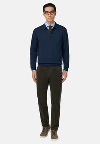 Marineblauer Pullover mit V-Ausschnitt und gerippten Bündchen, kombiniert mit olivgrünen Cordhosen und grauen Turnschuhen mit braunen Schnürsenkeln.