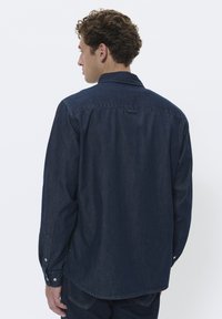 Camisa de mezclilla azul oscuro con cuello puntiagudo y mangas largas. Presenta puños abotonados y un dobladillo ligeramente curvado. Textura suave.
