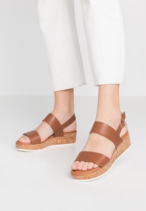 Pieds portant des sandales compensées en cuir marron avec plateforme en liège et semelle extérieure blanche, associées à un pantalon blanc court sur un fond clair.