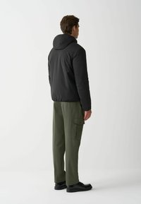 Schwarze Kapuzen-Puffjacke, locker sitzende olivgrüne Cargo-Hose mit seitlichen Taschen und schwarzen Schuhen, von hinten vor einem neutralen Hintergrund gezeigt.