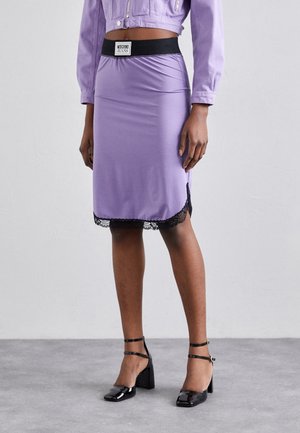 Kobieta ubrana w liliową ołówkową spódnicę z czarną koronkową lamówką, czarny elastyczny pas z napisem Moschino Jeans oraz czarne sandały na słupku z paskami.