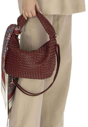 Ręka trzyma plecioną ciemnoczerwoną skórzaną torebkę z supełkowaną rączką i przymocowaną wzorzystą chustą, na tle beżowych spodni.