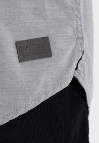 Tissu gris à texture lisse, avec un patch rectangulaire en cuir embossé du "19|98". Section en velours côtelé noir visible en bas.