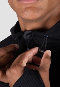 Schwarze Jacke mit hohem Kragen, die verstellbare Zugbänder und einen Reißverschluss hat. Der Stoff hat eine glatte Textur und ein tailliertes Design.