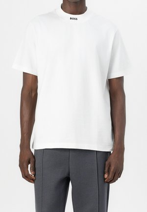 Mand iført en enkel hvid kortærmet skjorte med "BOSS" trykt ved kraven, kombineret med mørkegrå teksturerede bukser.