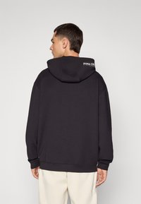 Sudadera con capucha negra de corte relajado, que presenta un logo en la parte posterior del cuello y puños acanalados. La tela parece suave con una textura lisa.