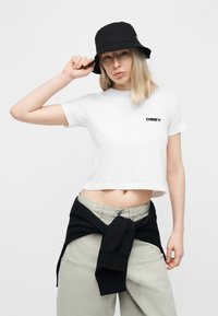 Γυναίκα που φοράει μαύρο καπέλο τύπου bucket, λευκό κοντομάνικο μπλουζάκι OBEY, ανοιχτόχρωμο παντελόνι και μαύρο φούτερ δεμένο γύρω από τη μέση της, με λευκό φόντο.