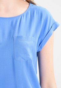 Modrá košile s krátkým rukávem vyrobená z lehkého materiálu, s malou kapsou vlevo, s ohrnutými rukávy a hladkou texturou.