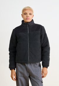 Veste en polaire noire avec col haut, dotée d'un empiècement en velours côtelé. Tissu texturé, fermeture éclair à l'avant et poignets côtelés. Portée avec un pantalon cargo gris.