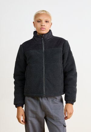 Veste en polaire noire avec col haut, dotée d'un empiècement en velours côtelé. Tissu texturé, fermeture éclair à l'avant et poignets côtelés. Portée avec un pantalon cargo gris.