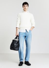 Cremefarbener Strickpullover, weißes Hemd, hellblaue Jeans und schwarze Lederschuhe. Hält eine strukturierte schwarze Tasche mit Reißverschluss und Tragegurt.