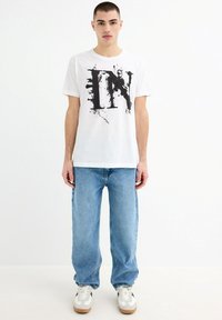 Camiseta de algodón blanca con un estampado gráfico negro, combinada con jeans de mezclilla azul claro y zapatillas gris y blancas. Corte relajado, estilo casual.