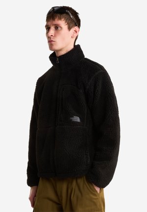 Jonge man met een zwarte fleece jas met rits en een bruine broek, met een zonnebril op zijn hoofd, staande tegen een effen achtergrond.