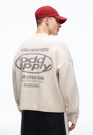 Mand iført cremet sweater med teksten "from nowhere odd supply for everyone" på ryggen og en rød kasket, stående sidevendt mod en hvid baggrund.