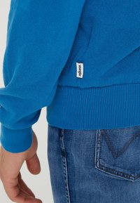 Niebieski sweter z dzianiny z prążkowanymi mankietami i dołem. Posiada małą białą metkę z napisem "Wrangler" z boku. Noszony z niebieskimi dżinsami.