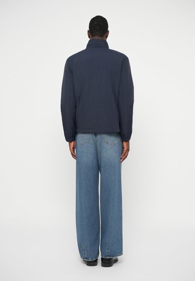 Άνδρας που φοράει σκούρο μπλε σακάκι και φαρδιά μπλε τζιν, στέκεται μετωπικά σε έναν λευκό τοίχο με τα χέρια του χαλαρά δίπλα του.