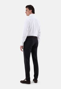 Camicia bianca con colletto rigido, abbinata a pantaloni scuri a righe sottili e mocassini in suede marroni, vista da dietro.