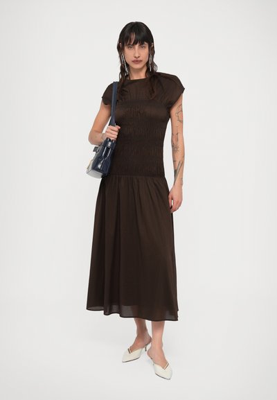 Γυναίκα με μεσάτο φόρεμα σε σκούρο καφέ χρώμα με πιετά, κρατώντας μια σκούρα μπλε τσάντα ώμου, φορώντας λευκά παπούτσια με χαμηλό τακούνι και μυτερή μύτη, στέκεται σε λευκό φόντο.