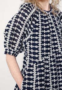 Femme portant une robe bleu marine avec des motifs géométriques en dentelle blanche et des manches courtes bouffantes, main reposant dans une poche latérale.