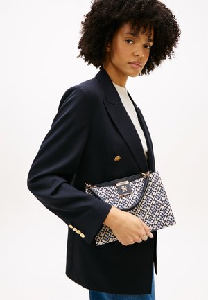Žena s kučeravými vlasmi, oblečená v čiernom blejzri so zlatými gombíkmi, drží béžovú a námornícky modrú vzorovanú kabelku so zlatým zapínaním.