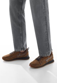 Persona che indossa sneakers in suede marroni con accenti neri e jeans grigio scuro, in piedi su una superficie bianca.