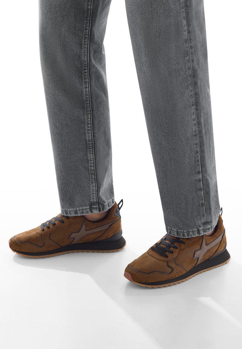 Persona che indossa sneakers in suede marroni con accenti neri e jeans grigio scuro, in piedi su una superficie bianca.