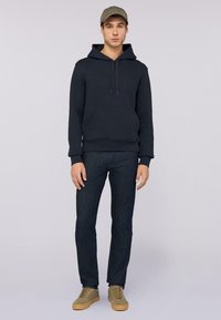Marinblå hoodie med framficka och snörning, kombinerad med mörka jeans och olivgröna skor. Modellen bär en matchande olivgrön keps.