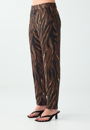 Pantalon marron et noir à rayures tigre porté avec des sandales noires à talons en forme de tongs sur une personne debout devant un fond uni.