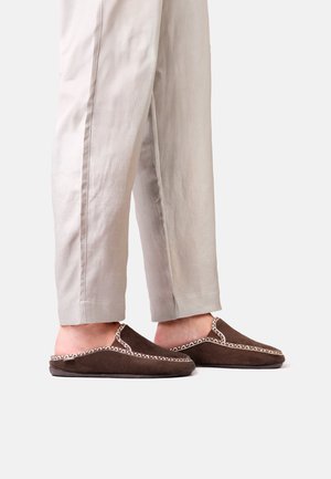 Personne portant un pantalon beige clair et des chaussons marron foncé à enfiler avec une légère bordure décorative, debout sur un fond clair uni.