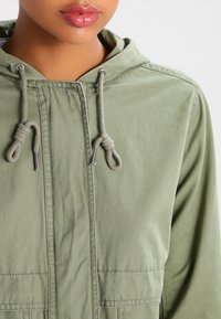 Olivengrøn hættejakke med snore, forlommer og et glat, let stof. Har metaldekorationer ved snoreenderne.