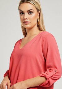 Blusa corallo con scollo a V, maniche a tre quarti e polsini con risvolto. Texture morbida con una vestibilità ampia e un leggero lucido.