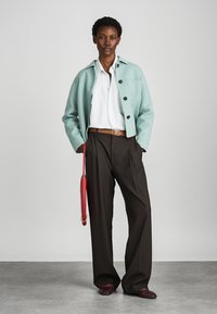 Παλτό σε μέντα απόχρωση, λευκό πουκάμισο, καφέ παντελόνι και κόκκινη τσάντα. Το μοντέλο φοράει μπορντό παπούτσια. Απλή φόντο, εστιασμένο στις λεπτομέρειες του outfit.